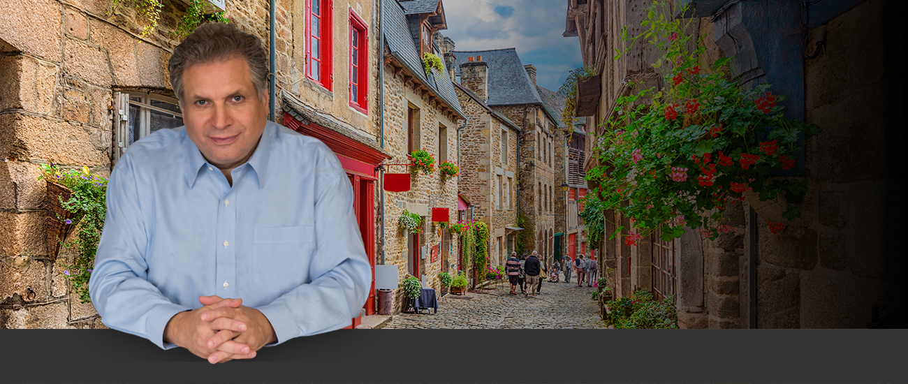 Frank Marciano in a light blue shirt is superimposed over a charming narrow street in a historic village, featuring stone houses with red and white shutters and abundant floral decorations.