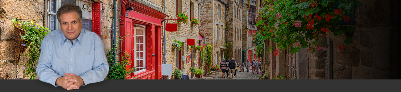 Frank Marciano in a light blue shirt is superimposed over a charming narrow street in a historic village, featuring stone houses with red and white shutters and abundant floral decorations.